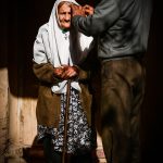 Photo of old woman getting her headscarf adjusted by a tall man with his back to the camera