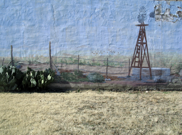 Photograph of a mural of a windmill painted on a wall above a yellowed grass field with a cactus.