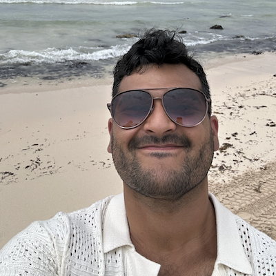 Headshot of Joe Lozano in a white shirt with sunglasses smiling against an ocean shore.