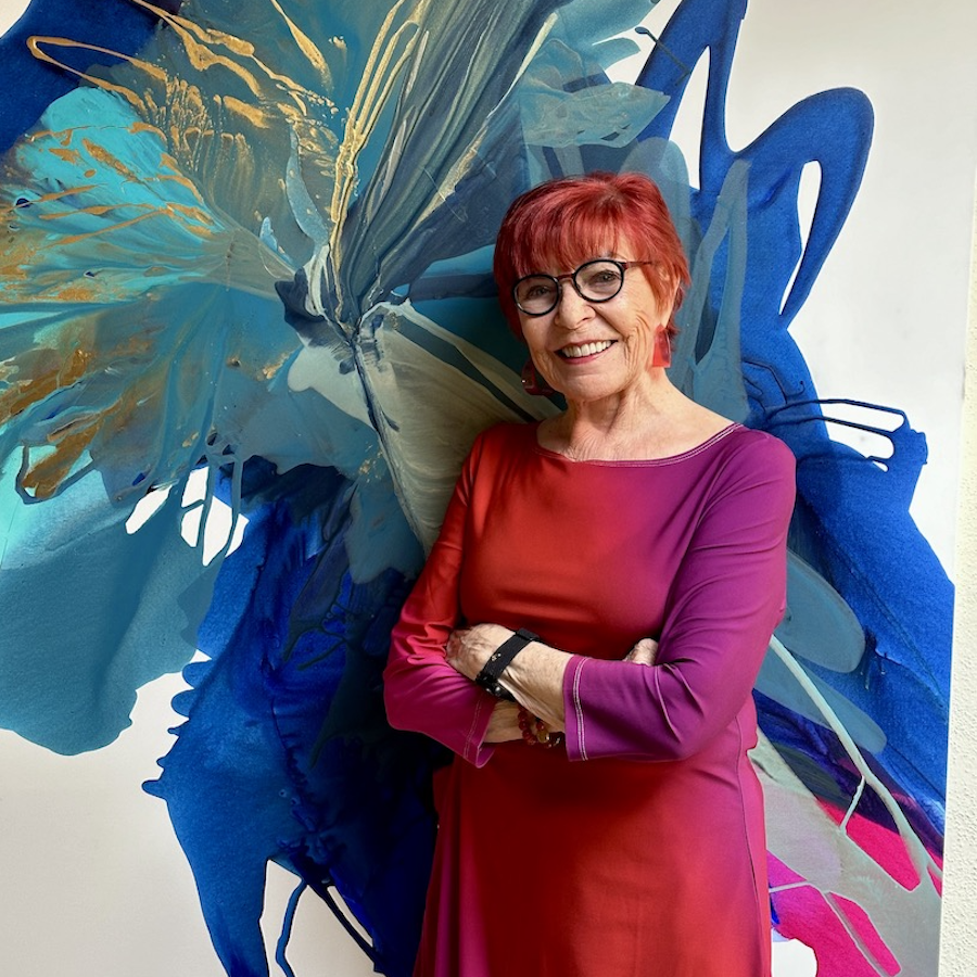 Headshot of Bette Ridgeway against one of her paintings in a red dress with matching red hair.