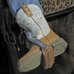 Photograph of a zoomed-in cowgirl boot, white and brown, in a stirrup against a brown horse's side, with a leopard-print saddle.