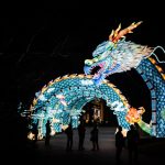 Photograph of neon-lighted blue dragon arch gate entrance with small human silhouettes walking below at night