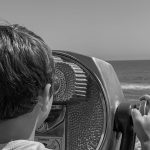 a photograph of a young boy looking through a binocular viewing stand at the beach and waves